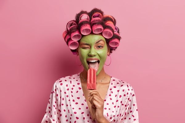 Divertente e allegra casalinga lecca un delizioso gelato, strizza l'occhio, si gode un rinfrescante dessert estivo freddo, indossa una maschera verde sul viso, bigodini, vestita con abiti informali, si diverte a casa