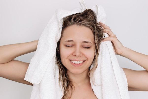 Una giovane donna sorridente che si asciuga i capelli bagnati con un asciugamano bianco sulla testa dopo la doccia.