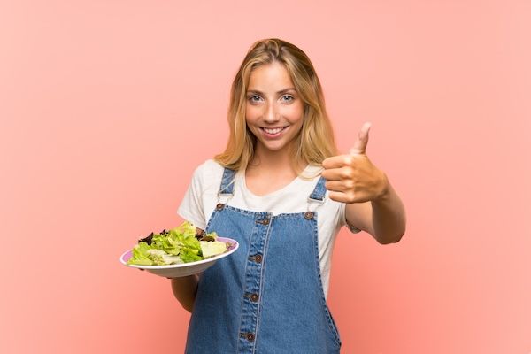 Giovane bionda con insalata su sfondo rosa, isolata con i pollici in su perché è successo qualcosa di buono
