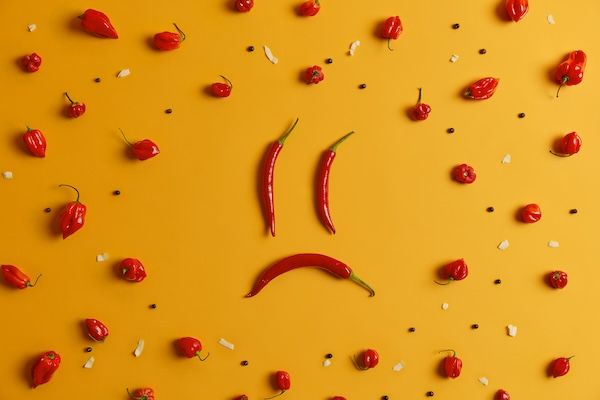 Volto umano triste fatto di peperoncino rosso lungo, peperone habanero con grani di pepe intorno, sfondo giallo. Scatto creativo, concetto di arte del cibo. Ingrediente piccante con nutrienti benefici. Vegetale piccante