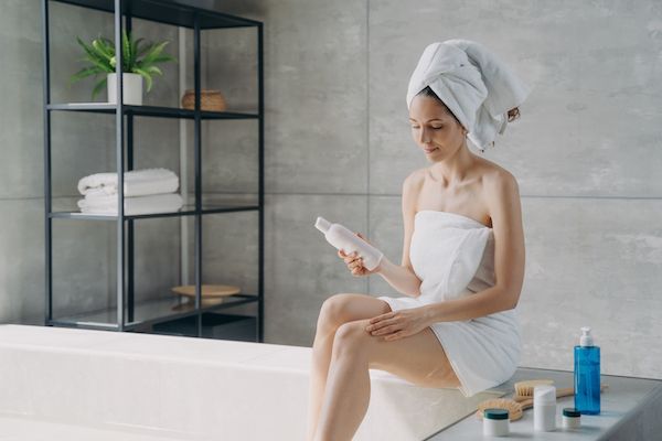 donna seduta in una vasca da bagno con panni sul corpo e sulla testa, con in mano un contenitore di crema che spalma sulla gamba