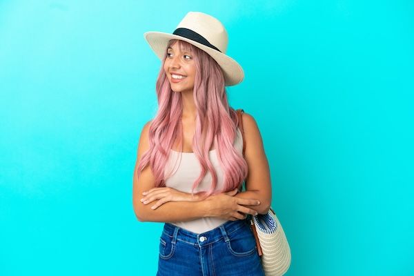 Giovane donna di razza mista che tiene una borsa da spiaggia con un cappello di paglia, isolata su sfondo blu, felice e sorridente.