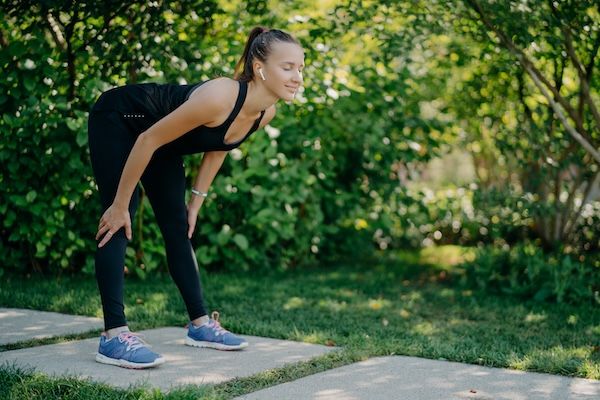 Donna fitness in abbigliamento sportivo nero tiene le mani sulle ginocchia, si inclina in avanti durante esercizi all'aperto contro la natura verde.