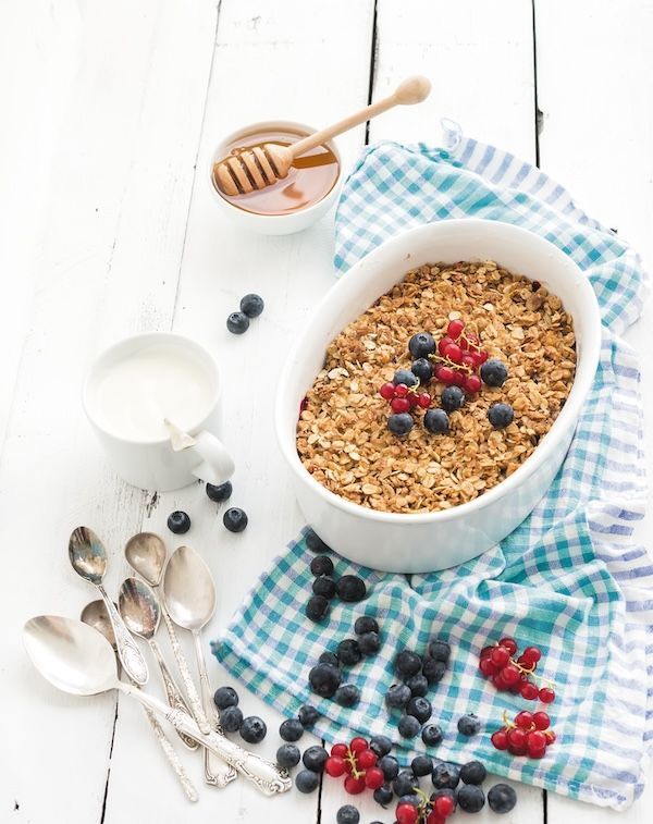 Una sana colazione con farina d'avena e muesli, crumble di frutti di bosco con yogurt fresco ai mirtilli e miele in una teglia in ceramica su uno sfondo rustico bianco