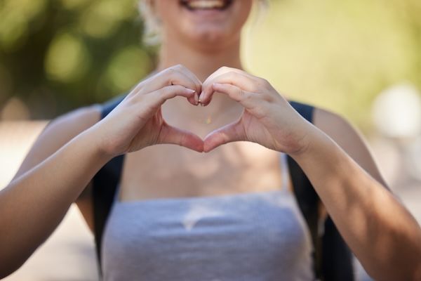Donna che si tiene per mano e ha un cuore emoji nella natura mentre è all'aperto per amore della libertà e sostegno per le avventure di viaggio estive Giovane donna felice con il segno della mano e uno zaino per motivare la pace e il benessere