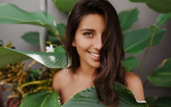 Bella signora con capelli ricci e occhi verdi sorride dolcemente Donna con ciglia lunghe in posa sullo sfondo di una pianta tropicale