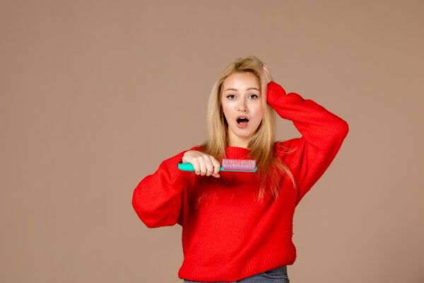 Ragazza bionda con maglione rosso in posa mentre si tocca i capelli mentre tiene una spazzola in mano con espressione sorpresa.